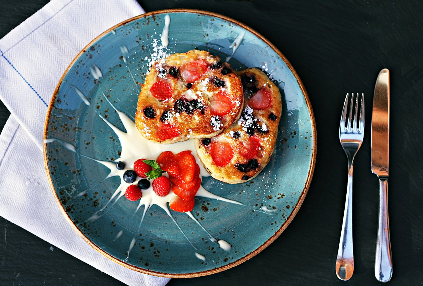 Two berry-topped pancakes on a blue plate with cutlery.