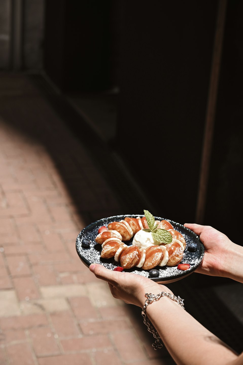 Hands hold a plate of mini pancakes with ice cream and berries.